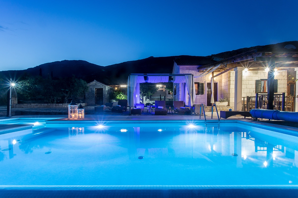 Image of the pool at night, with sun loungers and the villa in the background, dominated by the blue light from the pool.